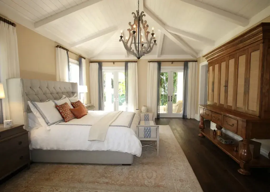 A spacious bedroom with a large bed, wooden furniture, chandelier, and light streaming in from French doors leading outside.
