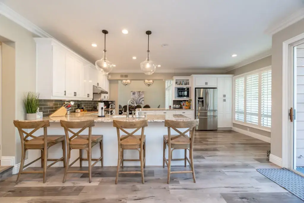 Modern kitchen with white cabinets, a marble island, four wooden barstools, stainless steel appliances, and pendant lights, with large windows letting in natural light.