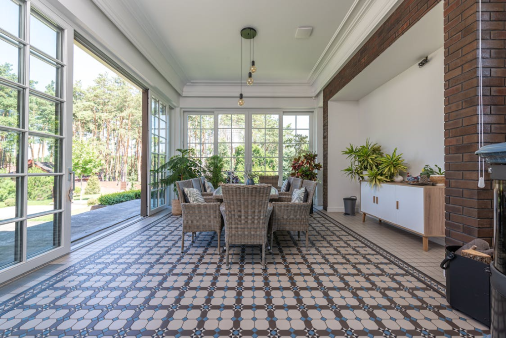A bright sunroom with large windows, wicker chairs around a table, patterned tile flooring, and various potted plants.