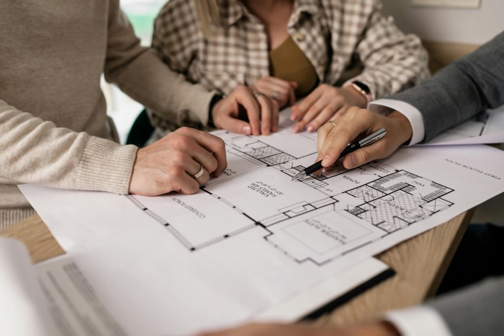 Three people review a floor plan blueprint together at a table, with one person pointing at the document with a pen.
