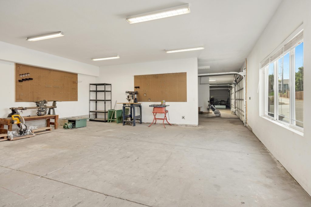 Spacious garage or workshop with pegboards on the walls, large windows, workbenches, metal shelves, and overhead fluorescent lighting.