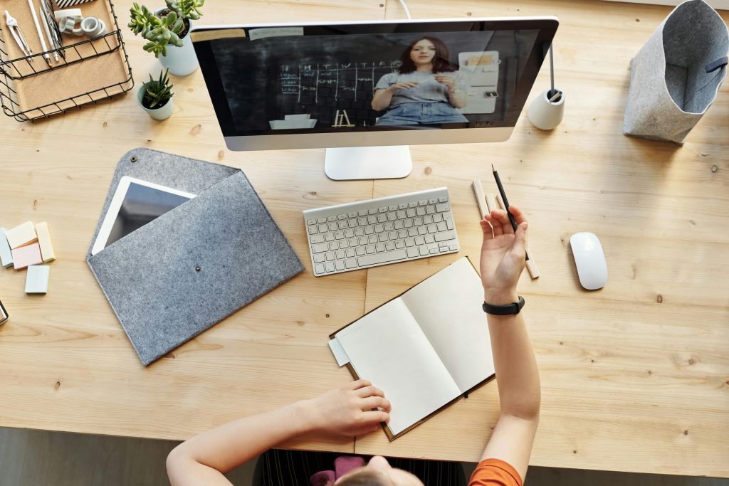 Person taking notes in a notebook while watching an online lesson on a desktop computer at a tidy wooden desk with various office supplies.