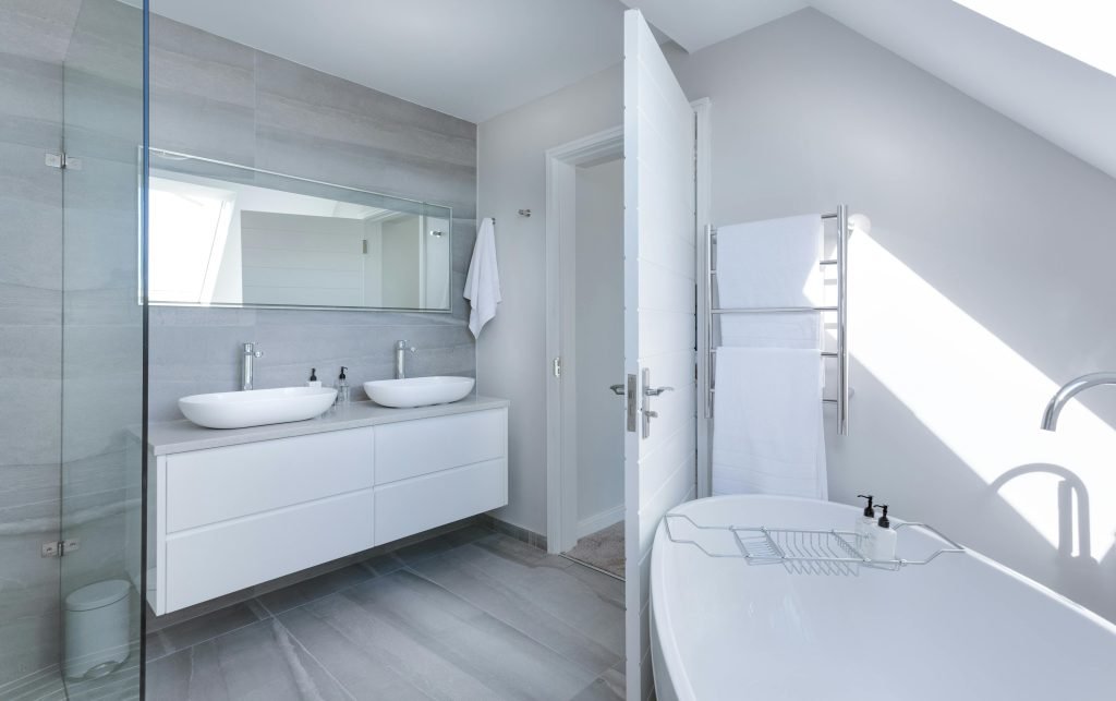Modern bathroom with two sinks, a large mirror, a white freestanding bathtub, a towel rack, and gray tiled walls and floor. Natural light enters from a skylight.