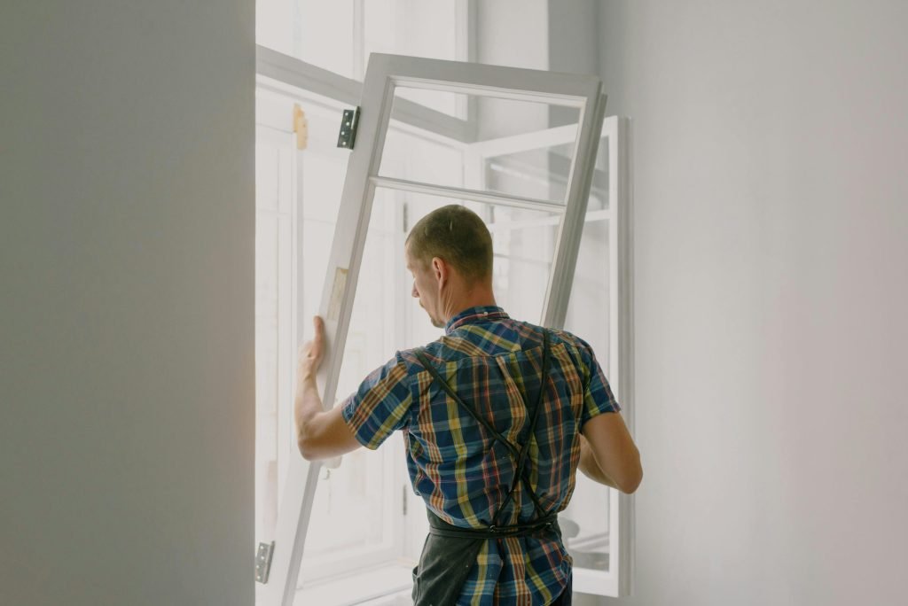 A person wearing an apron is installing or adjusting a white-framed window in a bright, minimally furnished room.