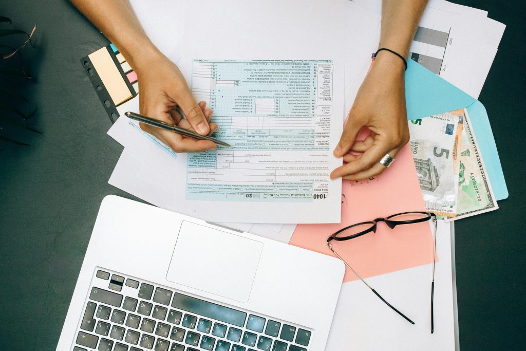 Person filling out a tax form at a desk with a laptop, eyeglasses, paper documents, and cash.