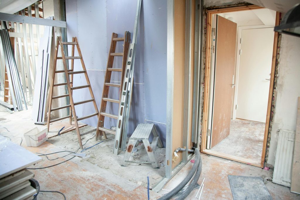 A room under renovation with ladders, construction materials, tools, and exposed pipes; a partially open door leads to another unfinished area.