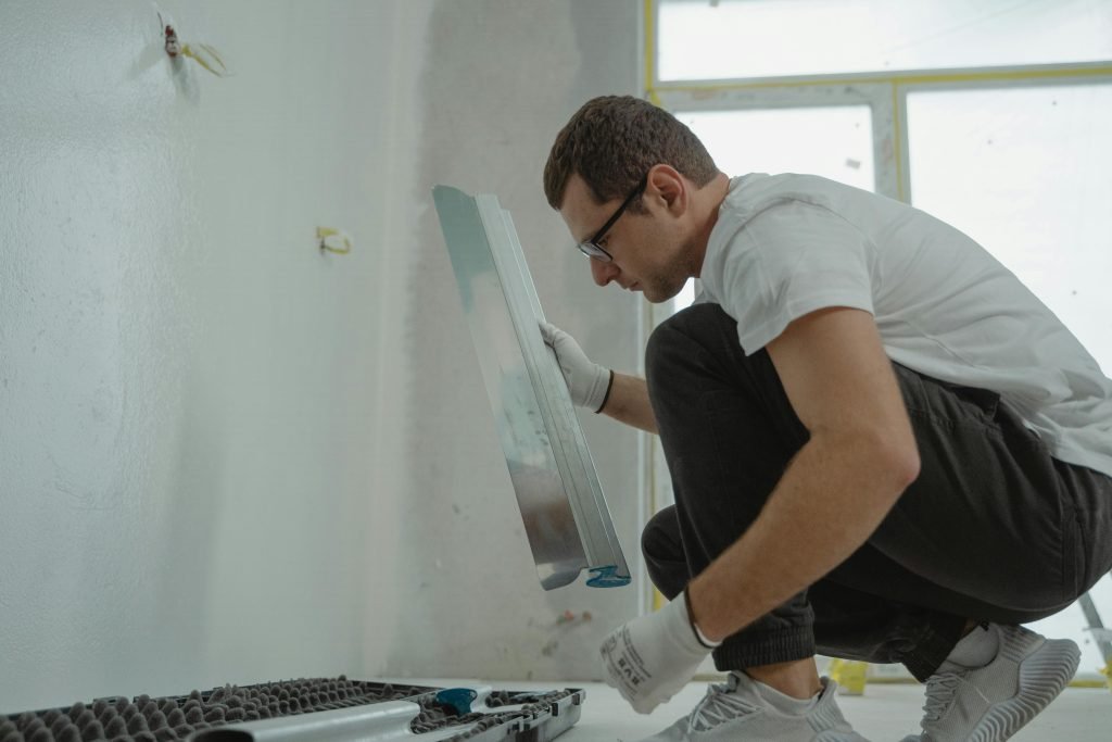 A man wearing glasses and gloves squats indoors, holding a large trowel near a wall, with painting tools and supplies on the floor beside him.